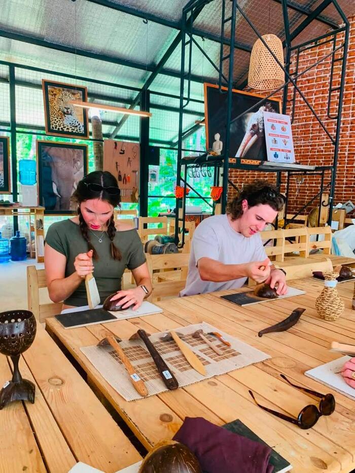 Two visitors shaping coconut shells with hand tools at the workbench during a coconut shell craft workshop in Sigiriya.
