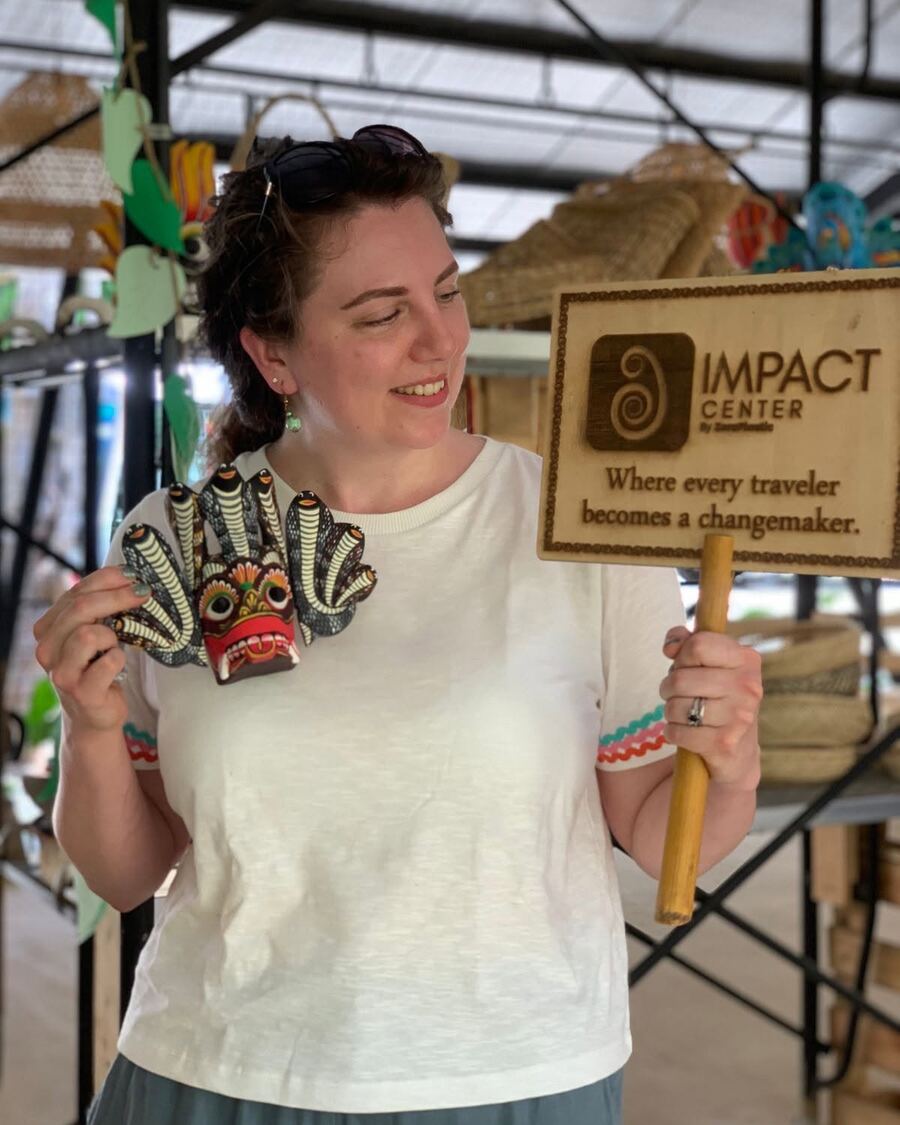 A visitor holding a carved Sri Lankan mask beside the Impact Center sign, part of the mask painting workshop in Sigiriya.