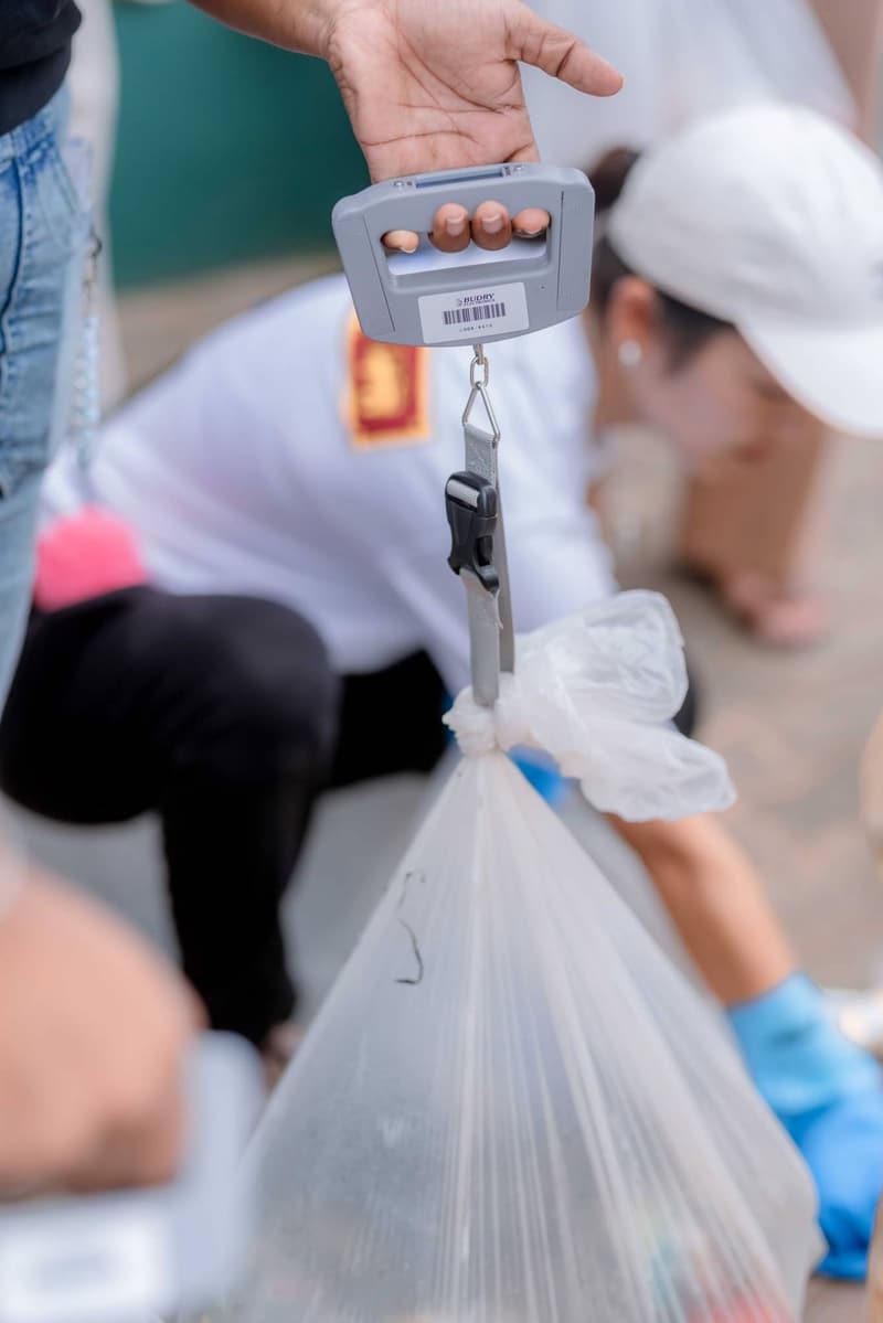 A hand holding a hanging scale with a clear bag of collected plastic waste attached, while a volunteer in a white cap and blue gloves gathers more plastic behind it.