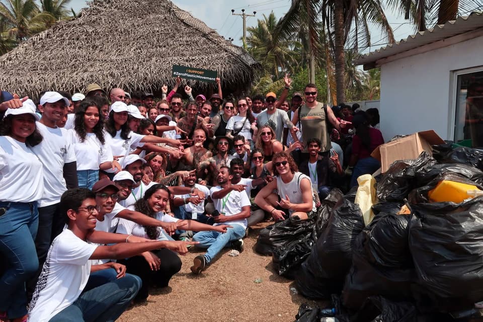 A large group of ZeroPlastic volunteers and international visitors posing together beside piles of black bags of collected plastic, one person holding an "I support #ZeroPlasticMovement" sign.