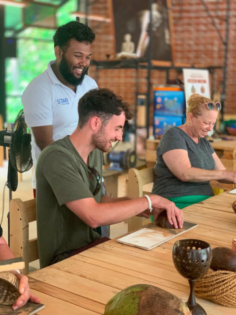 Smiling visitors sitting at a long wooden workbench trying a hands-on coconut-shell craft at the Impact Center in Sigiriya, while a team member looks on.