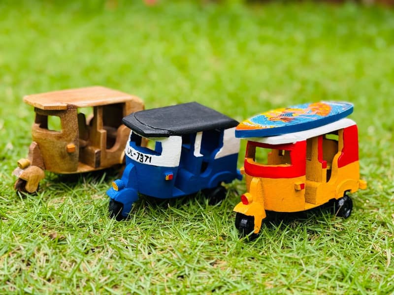 Three hand-carved wooden tuk-tuk models on grass, one unpainted and two finished in blue and yellow, made by Sri Lankan artisans.