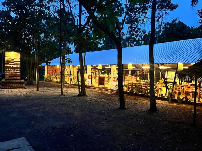 The ZeroPlastic Impact Center in Sigiriya lit up at dusk, an open timber and mesh pavilion among trees with an OPEN sign at the entrance.