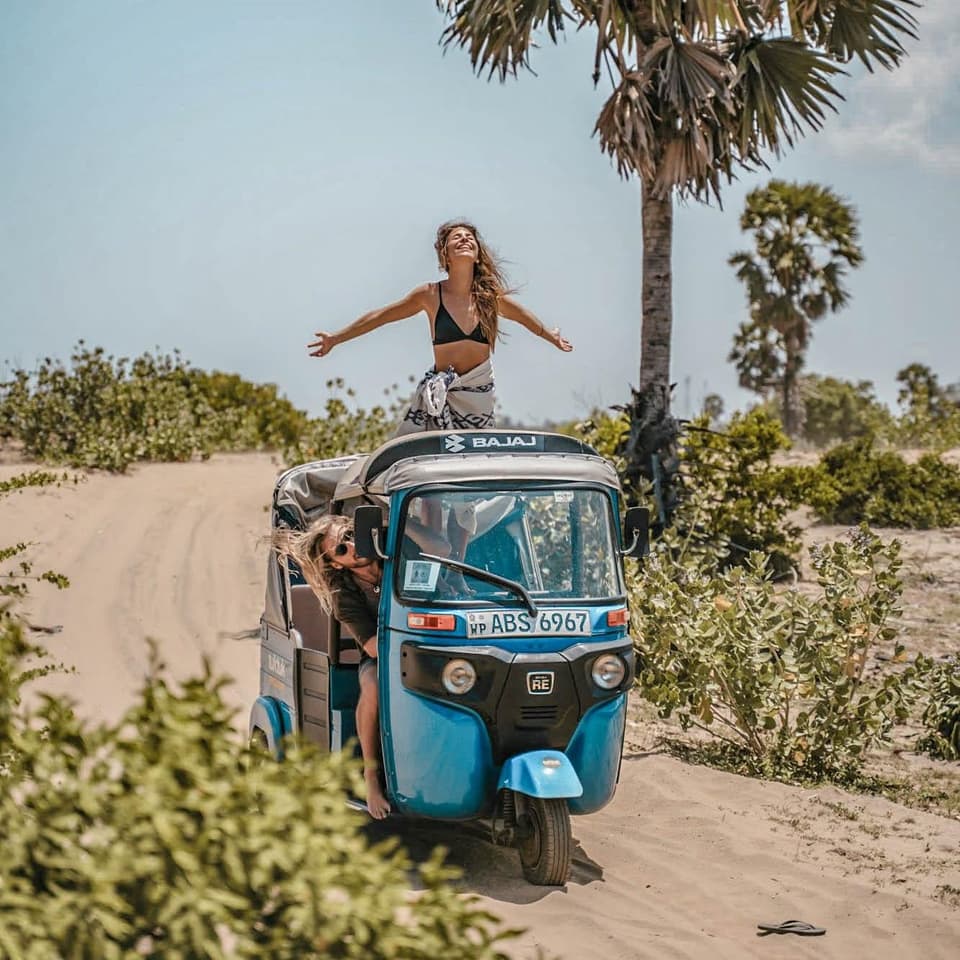 A traveller standing up through the roof of a tuk-tuk with arms outstretched on a sandy track lined with palms in Sri Lanka.