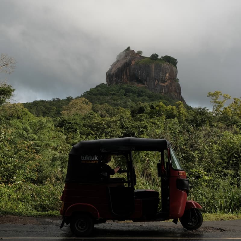 A hired tuk-tuk parked on the road below Sigiriya Lion Rock under a heavy sky.